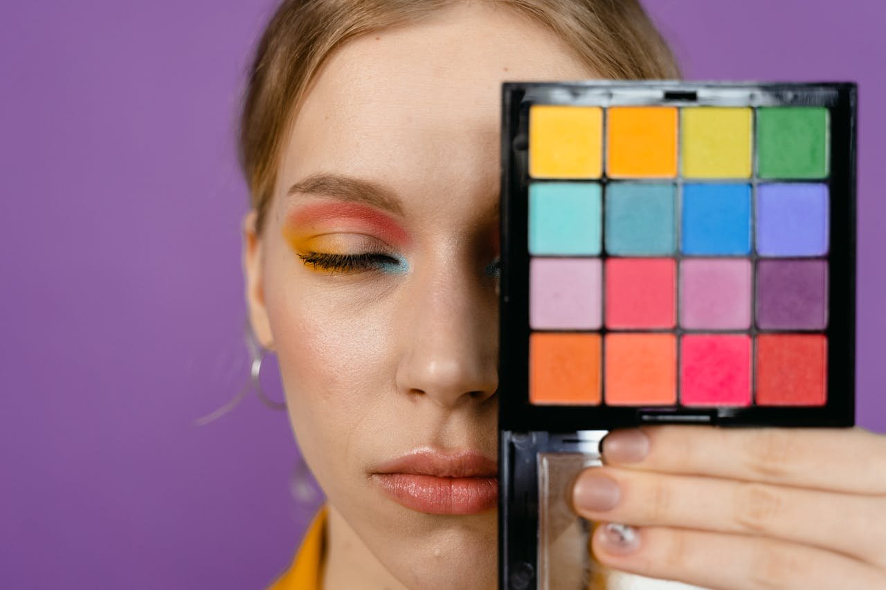 Close-up of a woman with colorful eyeshadow and palette against a purple background.